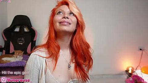 Media: Video of a smiling, fair-skinned woman with vibrant orange hair, wearing a white cardigan, in a cozy room with a gaming chair and a small table.