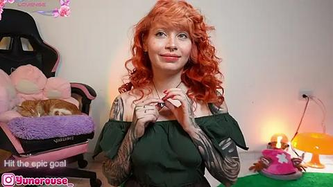 Media: Video of a pale-skinned woman with red curly hair, wearing a green off-the-shoulder top, standing in a room with a black gaming chair, plush toys, and a colorful lamp.