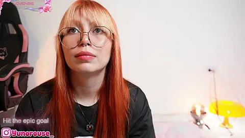 Media: Video of a fair-skinned, red-haired woman with glasses, wearing a black top, in a room with gaming equipment.