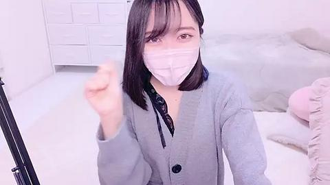 Media: A video of an East Asian woman with a light complexion, wearing a light gray cardigan and a white face mask, standing in a minimalistic white room.