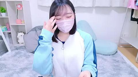 Media: Video of an Asian woman with straight black hair, wearing a blue and white hoodie, face mask, and glasses, adjusting her mask in a pastel-themed room with plush toys and white furniture.