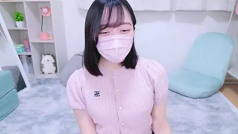 Media: Video of a young Asian woman with straight black hair and light skin, wearing a pink face mask and button-up shirt, seated on a light grey carpeted floor in a minimalist room with light blue furniture and white walls.