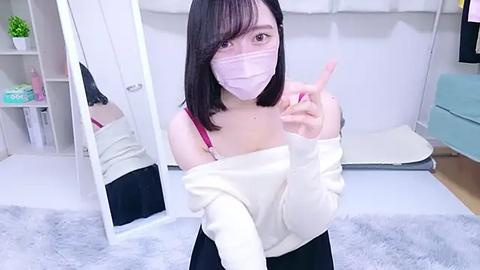 Media: Video of an East Asian woman with straight black hair, wearing a pink mask, off-shoulder white sweater, and black skirt, standing in a modern, minimalist bedroom with white walls, gray carpet, and white shelves.