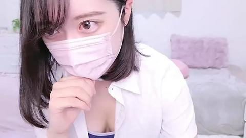 Media: Video of an Asian woman with shoulder-length dark hair, wearing a white shirt and a face mask, covering her mouth. She holds a piece of paper near her mouth. Background features a white wall and a bed with light-colored sheets.