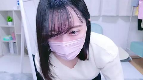 Media: Video of an Asian woman with long black hair and bangs, wearing a white mask, standing in a clean, white-tiled room with shelves and a blue chair.