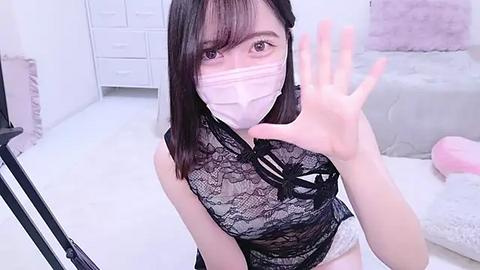 Media: Video of a young Asian woman in a white mask, black lace bodysuit, and white panties, waving from a plush, pastel-colored room with white furniture and a furry toy.