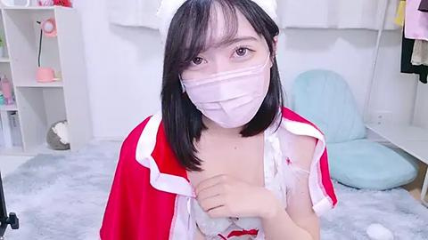 Media: Video of an East Asian woman in a red and white maid outfit, wearing a face mask, with shoulder-length black hair, standing in a bright, modern room with white walls, light blue pillows, and a fluffy rug.