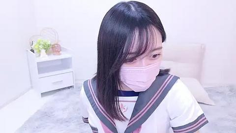 Media: Video of an Asian woman in a schoolgirl uniform with pink face mask, standing on a white carpet in a white room with a nightstand and plant.