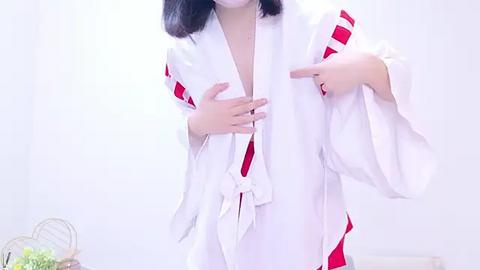 Media: Video of an East Asian woman with shoulder-length black hair wearing a white, open-front robe with red trim, pointing at her chest. The background is plain white.