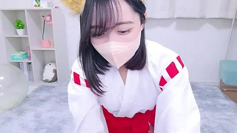 Media: Video of an Asian woman with short black hair, wearing a white kimono with red accents, a surgical mask, and a fluffy white hat, sitting on a grey rug in a brightly lit room with white walls and shelves.