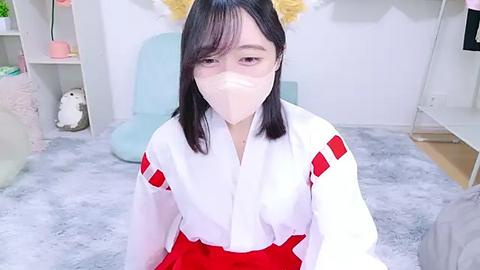 Media: Video of an East Asian woman in a white hakama with red accents, wearing a white mask, sitting on a plush carpet in a light-filled room with pastel furniture and soft decor.