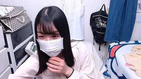 Media: Video of an East Asian woman with long black hair and a white mask, sitting on a bed with a blue blanket, in a white room with laundry baskets, a black bag, and a blue curtain.