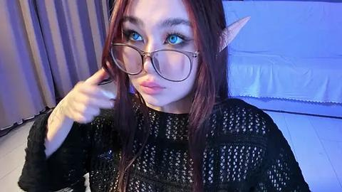 Media: Video of a young woman with fair skin, long brown hair, and blue eyes. She wears large, round glasses and pointy elf ears. She is dressed in a black, textured sweater. Background features a white bed and beige curtains.