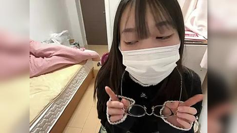 Media: A video of an Asian woman with straight black hair and bangs, wearing a white mask and black top, pointing at something, standing in a cluttered room with a bed and pink sheets.