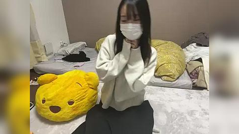 Media: A video of a young Asian woman with long black hair, wearing a white mask and a cream sweater, sitting on a messy bed with a yellow teddy bear and yellow pillows.