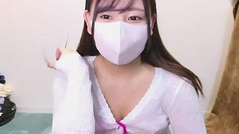 Media: Video of an Asian woman with light skin, wearing a white face mask, white long-sleeve top, and brown hair. She's indoors, standing with her right hand on her shoulder, in a casual setting.