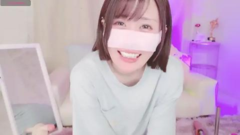 Media: A video of a smiling, light-skinned woman with a bob haircut and light blue eyes, wearing a white cardboard box as a virtual reality headset, in a softly lit, pastel-toned room with a white couch and a shelf with trophies.