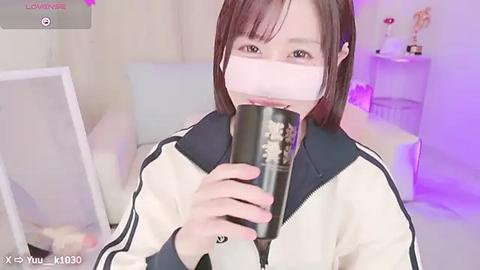 Media: A video of an Asian woman with straight brown hair, wearing a face mask, drinking from a black cup, in a room with white walls, a white couch, and a shelf with trophies.