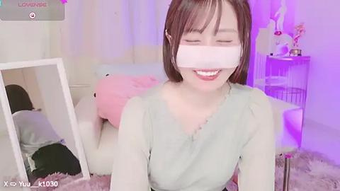 Media: Video of an Asian woman with shoulder-length brown hair, smiling, wearing a VR headset, a light green shirt, and sitting on a plush rug. Background features a white bed, pink stuffed toy, and purple lighting.