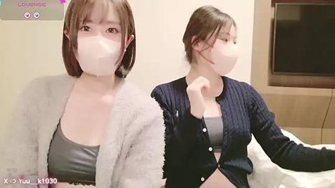 Media: Video of two young Asian women with short brown hair, wearing white face masks, grey tops, and dark cardigans. One poses seductively on a bed, while the other sits behind her, both in a dimly lit room.