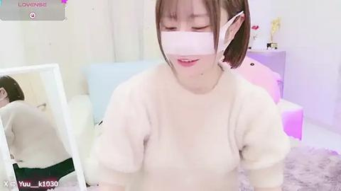 Media: Video of a young Asian woman with short brown hair wearing a white mask and a cozy sweater, seated in a softly lit room with pastel furniture and a plush rug.