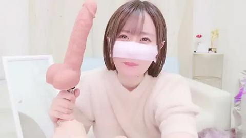 Media: Video of an Asian woman with short brown hair, wearing a pink surgical mask, holding a large erect penis, in a bright, sterile room with white walls and minimal decor.