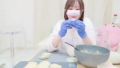 Media: A video of a young Asian woman in a white lab coat and blue gloves meticulously preparing dough on a kitchen counter. A grey mixing bowl with dough and egg yolk is in the foreground, and the background features soft, pastel-colored decor.