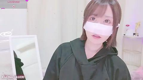 Media: A video of a young woman with straight brown hair, wearing a black hoodie and a white surgical mask, in a light-colored room with a white cabinet and a pink bed.