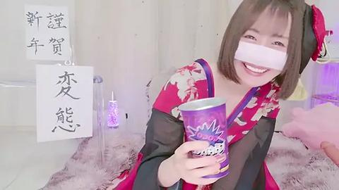 Media: Video of a smiling East Asian woman in a red kimono with a black floral pattern, holding a purple cup with Japanese characters, in a softly lit room with white curtains and pink fur rug.