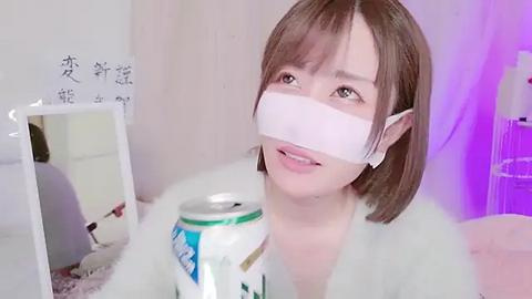 Media: A video of a young woman with short brown hair, wearing a white face mask and a fluffy white bathrobe, holding a can of soda. The room is softly lit with purple and white lights.