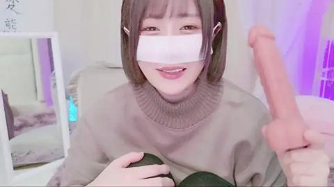 Media: Video of an East Asian woman with short brown hair, wearing a face mask, beige sweater, and holding an erect penis, indoors with a mirror and purple lighting.