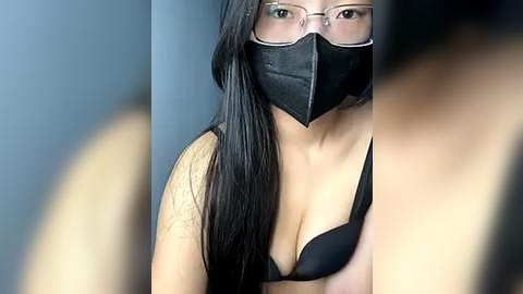 Media: Video of a woman with long black hair, wearing a black mask, glasses, and a black bra, with a blurred background.