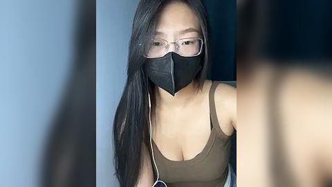 Media: Video of an East Asian woman with long black hair, wearing glasses, a black face mask, and a tan tank top. She appears to be indoors, possibly a medical setting.