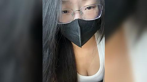 Media: Video of an Asian woman with long black hair, wearing clear glasses and a black surgical mask, in a white tank top, looking down. Background is blurred.