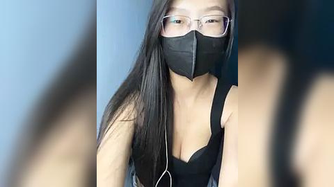 Media: A video of an East Asian woman with long black hair, wearing a black face mask, black glasses, and a black tank top. She's seated indoors, with blurred out-of-focus figures in the background.