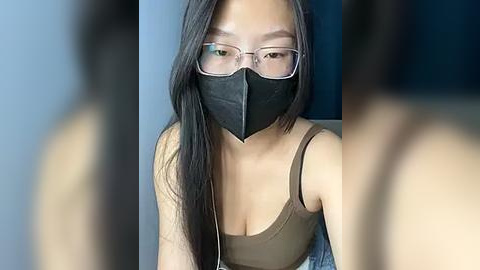 Media: Video of an Asian woman with long black hair, wearing glasses, a black mask, and a brown tank top, sitting indoors.