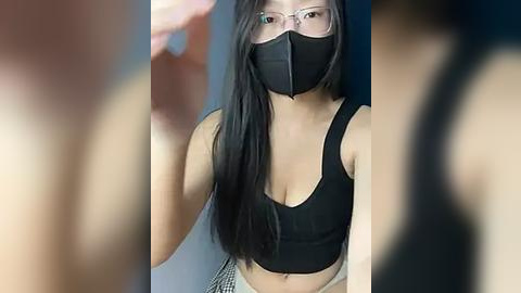 Media: Video of an Asian woman with long black hair, wearing a black face mask, black tank top, and checkered skirt. Blurry background suggests indoor setting.