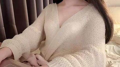 Media: Video of a light-skinned woman with long, straight brown hair, wearing a cream-colored, textured, V-neck cardigan over a white shirt, sitting on a bed with a beige blanket, against a backdrop of dark brown curtains.