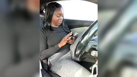 Media: A video of a woman with medium-dark skin and straight black hair, wearing a black long-sleeved top and grey patterned pants, attentively texting on her phone while sitting in a black leather car seat.