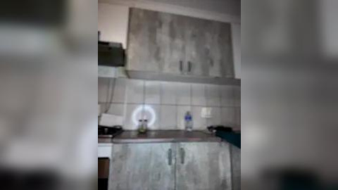 Media: A blurry video of a small, outdated kitchen with light gray cabinets, white tiled backsplash, and minimal counter space, featuring a white fan, a blue water bottle, and a black appliance.