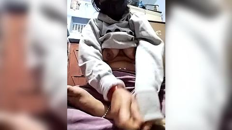 Media: Video of a woman with light skin, wearing a grey hoodie and white pants, lifting her hoodie to reveal her bare breasts. She sits on a bed with a pink blanket, hands touching her feet. Background includes a white cabinet and a fan.