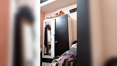 Media: A video of a cluttered room with a black and white mirror wardrobe, a small table, and a bed with a purple and white patterned blanket. The room has peach-colored walls and a narrow doorway in the background.