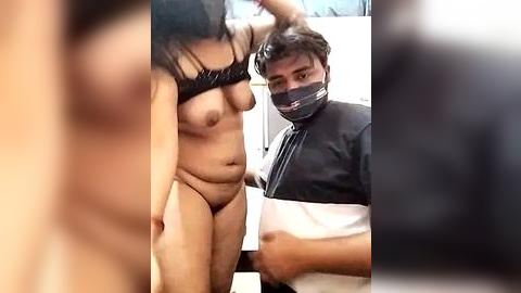 Media: Video of a naked woman with medium-sized breasts, bound wrists, and a cloth over her eyes, kneeling on a bed; a man in a black mask and shirt holds her head.