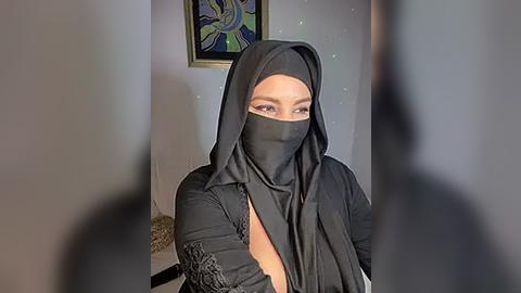 Media: Video of a woman in a black niqab and headscarf, seated indoors, with a framed abstract art piece in the background.