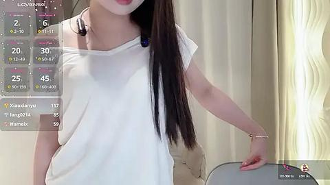 Media: Video of an Asian woman with long, straight black hair wearing a white off-shoulder blouse, standing indoors. Background includes a cream-colored curtain and a digital health tracker display.