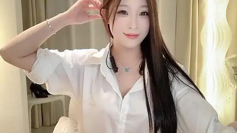 Media: A video of an East Asian woman with long, straight black hair, wearing a white blouse, smiling, adjusting her hair, in a softly lit room with beige curtains.