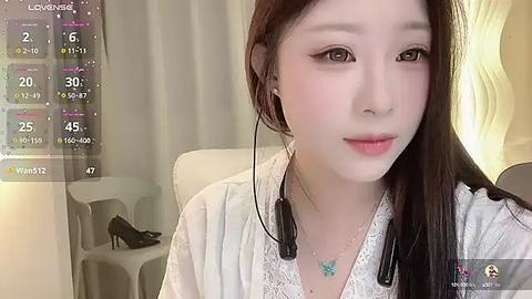 Media: Video of an Asian woman with fair skin and long dark hair, wearing headphones and a white lace top, sitting indoors with a white chair and a lamp in the background.