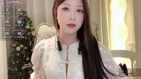 Media: Video of an East Asian woman with long brown hair, wearing a white lace blouse, standing in front of a decorated Christmas tree in a warmly lit room.