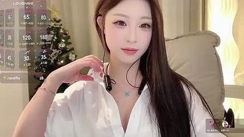 Media: Video of an East Asian woman with long black hair, wearing a white shirt, sitting indoors. Background shows a cozy room with a lit tree and soft lighting.