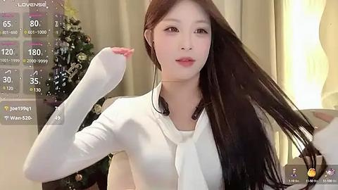 Media: Video of an East Asian woman with long, straight black hair, fair skin, wearing a white long-sleeved top, posing indoors in front of a flower arrangement, with a live video overlay showing weather data.